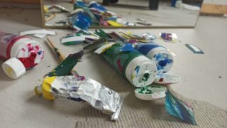 Mehrere Farbtuben, ein Pinsel und Stücke einer zerrissenen Farbverpackung liegen verstreut auf dem Boden vor einem Spiegel.