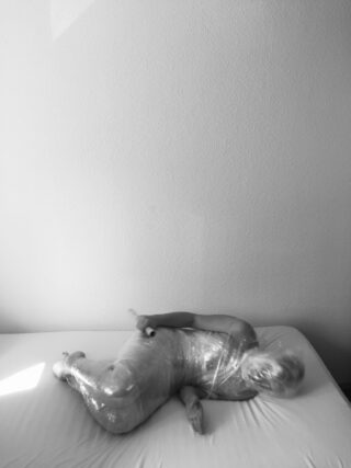 Eine Person liegt auf einer Matratze, die fest in durchsichtigen Kunststoff eingewickelt ist. Kopf und Körper sind sichtbar. Sie lehnt an einer kahlen Wand in einem spärlich möblierten Raum.