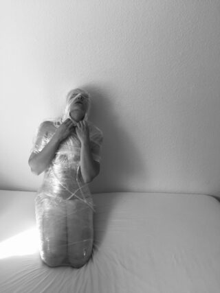 Auf einer Schwarzweißfotografie kniet eine Person, die in Plastikfolie eingewickelt ist, die Hände in der Nähe ihres Halses und blickt nach oben auf eine schlichte Wand.