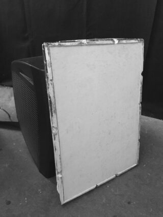 Ein großer Röhrenfernseher mit einem rechteckigen weißen Panel, das an seinem Bildschirm lehnt, auf einem Betonboden mit dunklem Stoffhintergrund.