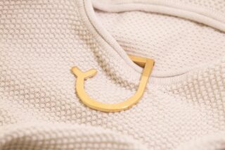 Ein goldfarbener Metallhaken in Form eines umgedrehten Buchstabens „J“ ruht auf einem strukturierten cremefarbenen Stoff.