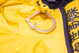 Nahaufnahme einer gelben Jacke mit einem goldenen Metallkarabiner, der in der Nähe eines Reißverschlusses mit schwarz-gelbem Text befestigt ist.