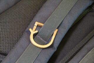 Ein goldener Metallhaken, der an einem grünen Stoffriemen an einem dunkelfarbigen Rucksack befestigt ist.