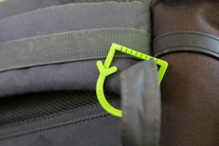 Nahaufnahme eines grünen Plastikhakens, der am Riemen eines schwarzen Stoffrucksacks befestigt ist.