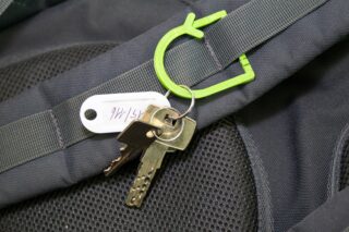 Ein Schlüsselsatz, der an einem grünen Karabinerhaken an einem grauen Rucksackgurt befestigt ist, einschließlich eines weißen Schlüsselanhängers mit der Aufschrift „151 4W4“.