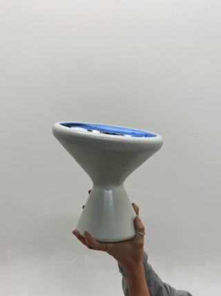 Eine Hand hält eine weiße Keramikvase mit einem breiten, flachen blauen Inneren vor einem schlichten hellgrauen Hintergrund hoch.