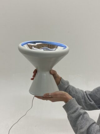 Eine Person in einem grauen Sweatshirt hält eine moderne, kegelförmige weiße Lampe mit einer reflektierenden Schale und blauem Rand. Das Kabel der Lampe ist sichtbar.