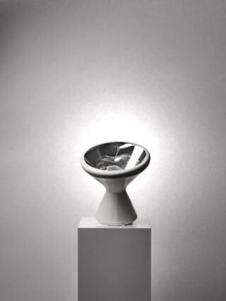 Ein modernes, metallisches, schalenförmiges Objekt mit konischer Basis wird auf einem weißen Sockel vor einem schlichten, hellgrauen Hintergrund präsentiert.