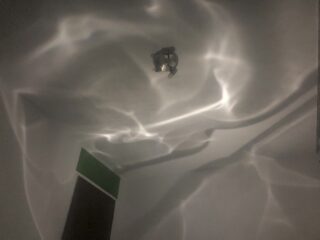 Eine Deckenleuchte wirft wellenförmige, abstrakte Schatten und Reflexionen auf eine weiße Decke über einem grün-braunen rechteckigen Objekt.