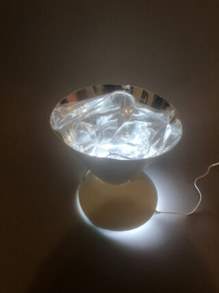 Eine moderne, weiße Tischlampe mit einem reflektierenden, zerknittert wirkenden Schirm, der helles weißes Licht ausstrahlt und auf einer dunklen Oberfläche mit sichtbarem Netzkabel platziert ist.