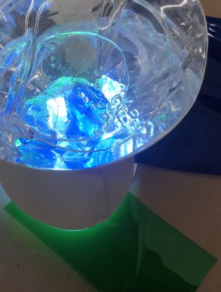 Eine wirbelnde klare Flüssigkeit in einer durchsichtigen Schüssel wird von blauem und grünem Licht beleuchtet, das farbige Reflexionen auf einen weißen Untergrund und nahe gelegene Oberflächen wirft.