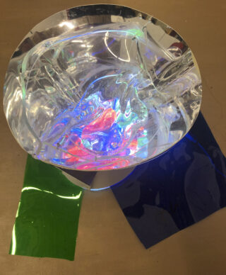 Eine reflektierende, zerknitterte transparente Schüssel mit buntem Licht im Inneren, neben grünen und dunkelblauen glänzenden Plastikfolien auf einer braunen Oberfläche.