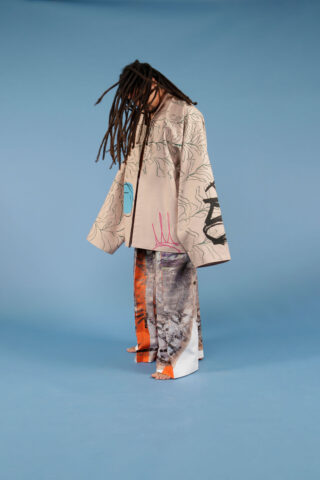 Person mit langen Dreadlocks trägt eine gemusterte beige Jacke und weite Hosen mit abstrakten orangefarbenen und grauen Aufdrucken und steht barfuß vor einem blauen Hintergrund.