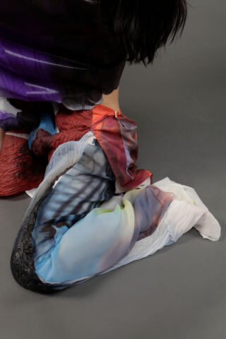 Eine Person kniet auf einer grauen Fläche. Sie ist in einen mehrfarbigen, strukturierten Stoff in den Farben Rot, Blau, Weiß, Schwarz und Lila gekleidet. Nur ihr Unterkörper ist sichtbar.