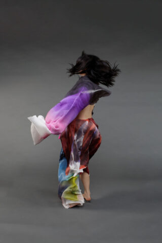 Vor einem schlichten grauen Hintergrund wird eine Person mit wehendem Haar und farbenfroher, wallender Kleidung mitten in der Drehung aufgenommen.
