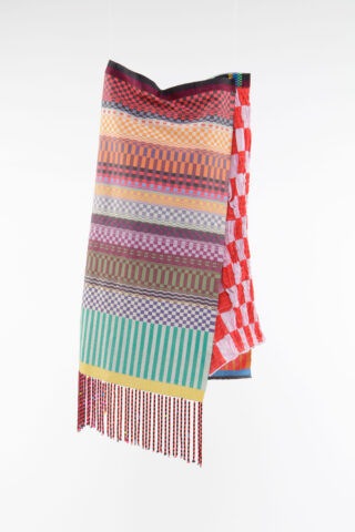Vor einem weißen Hintergrund hängt ein bunt gewebter Stoff mit verschiedenen Schachbrettmustern und Streifen in Rot, Gelb, Grün, Lila und Orange, versehen mit einem Fransenrand.