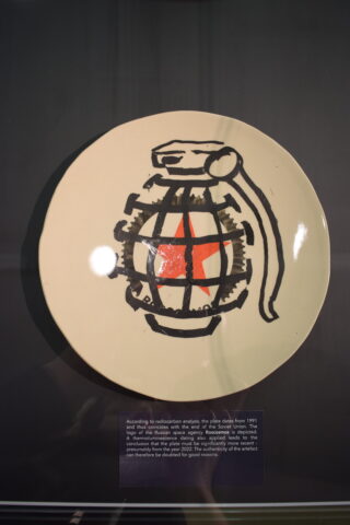 Ein Keramikteller mit der gemalten Abbildung einer Handgranate mit einem roten Stern in der Mitte, hinter Glas ausgestellt, darunter befindet sich eine Informationstafel.
