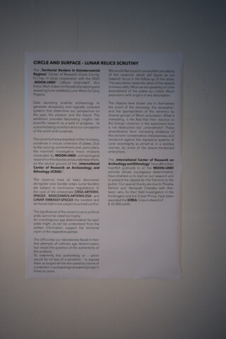An einer weißen Wand ist ein gedrucktes Dokument mit dem Titel „Kreis und Oberfläche – Untersuchung von Mondrelikten“ befestigt, das Text über archäologische Studien und Mondobjekte enthält.