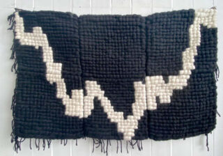 Ein rechteckiger gewebter Stoff mit schwarzem Hintergrund und einer gezackten weißen Linie, die horizontal durch die Mitte verläuft und an einer weißen Wand hängt.