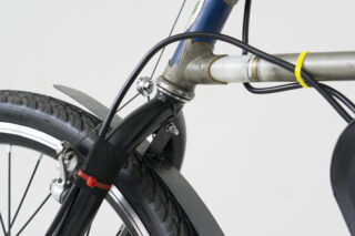 Nahaufnahme des Vorderrads eines Fahrrads. Zu sehen sind der schwarze Reifen, die Gabel, das Schutzblech, die Bremskabel und ein Teil des Rahmens vor einem schlichten Hintergrund.