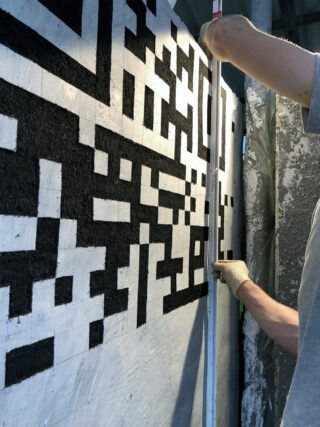 Eine Person misst mit einem Maßband eine Wand, die mit einem schwarz-weißen geometrischen Muster bemalt ist.