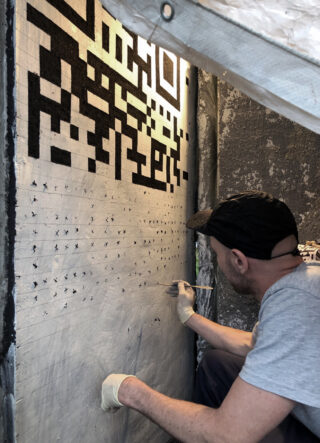 Eine Person mit Handschuhen und Mütze malt mit einem Pinsel schwarze geometrische Formen auf eine mit X-förmigen Markierungen bedeckte Wand.