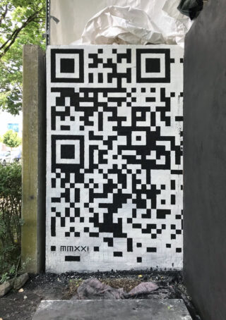 Auf eine Außenwand ist ein großer schwarz-weißer QR-Code gemalt. In der unteren linken Ecke stehen die römischen Ziffern „MMXXI“.