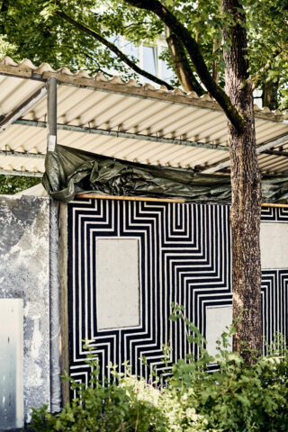 Eine Betonwand weist unter einem Wellblechdach ein kräftiges schwarz-weißes geometrisches Muster auf, im Vordergrund sind ein Baum und Grünpflanzen zu sehen.