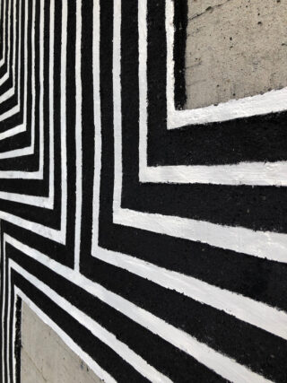 Nahaufnahme einer Betonwand, die mit sich kreuzenden parallelen schwarzen und weißen Linien bemalt ist, die geometrische Muster bilden.