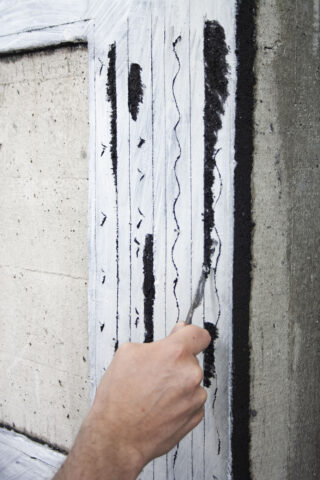 Eine Hand trägt mit einem Werkzeug schwarzes Material entlang wellenförmiger und gerader vertikaler Linien auf einer weiß gestrichenen Betonoberfläche auf oder kratzt es ab.