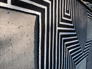 Eine Betonwand mit schwarz-weißen geometrischen Streifen, die um zwei rechteckige Abschnitte aus Sichtbeton gemalt sind.