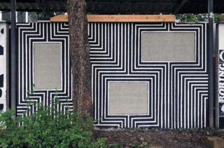 Eine Wand mit schwarz-weißen geometrischen Linienmustern und drei rechteckigen grauen Abschnitten, die teilweise von einem Baum und Pflanzen im Vordergrund verdeckt werden.