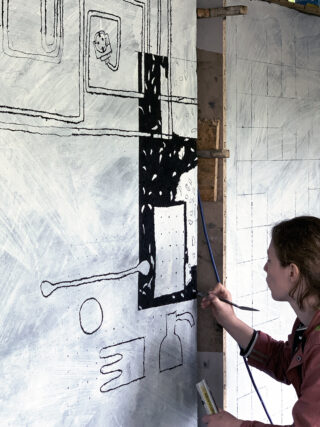 Eine Person malt schwarze geometrische und abstrakte Formen auf eine große, teilweise skizzierte Leinwand, die an einer Wand hängt.