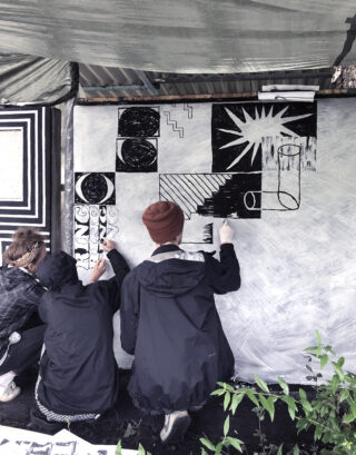 Drei Personen in Jacken malen unter einer Plane ein großes, abstraktes Schwarz-Weiß-Wandbild auf eine Leinwand im Freien.