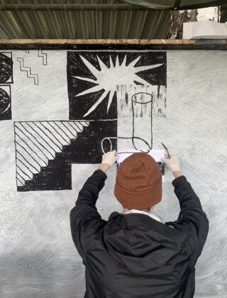 Eine Person mit einer braunen Mütze und einer schwarzen Jacke hält Papier an eine Wand, während sie geometrische Formen und Muster auf ein Wandgemälde zeichnet.