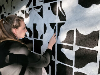 Eine Person verwendet einen Pinsel oder ein Werkzeug, um ein abstraktes Schwarzweiß-Wandbild auf einer strukturierten Wand zu malen oder auszubessern.