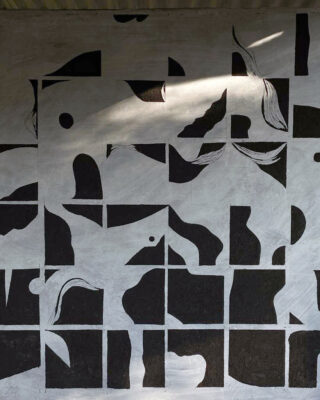 Ein geometrisches Wandbild mit abstrakten Formen in Schwarz und Weiß, aufgeteilt in ein Raster aus Rechtecken, wird von einem Sonnenstrahl von oben beleuchtet.