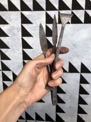 Eine Hand, die drei Bildhauerwerkzeuge aus Metall vor einem schwarz-weißen, geometrisch gemusterten Hintergrund hält.