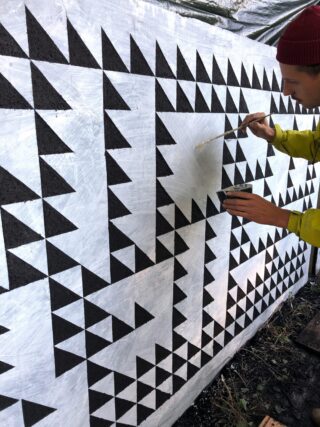 Eine Person mit einer roten Mütze und einer gelben Jacke malt ein geometrisches Wandbild mit schwarzen Dreiecken auf eine weiße Wand.