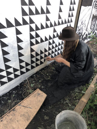 Eine Person mit dunklem Hut und Mantel kniet und malt ein schwarz-weißes geometrisches Dreiecksmuster auf eine Außenwand.