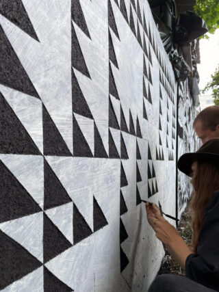 Zwei Personen malen ein Wandbild aus schwarz-weißen geometrischen Dreiecken auf eine Außenwand.
