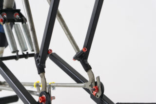 Nahaufnahme einer mechanischen Struktur mit Metallstangen, schwarzen Stützen, roten Bolzen und Spiralfedern vor einem schlichten weißen Hintergrund.