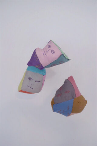 Zwei bunte, unregelmäßig geformte Stoffobjekte mit handgezeichneten Gesichtern und Augen schweben vor einem schlichten weißen Hintergrund.