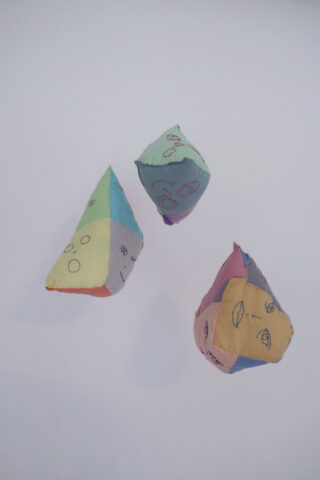 Drei bunte, handgenähte Stoffformen mit einfach gestickten Gesichtern und Symbolen sind auf einem schlichten weißen Hintergrund angeordnet.