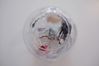 Eine durchsichtige Schüssel mit verschiedenen kleinen Objekten wie Papier, Schnur und Plastikstücken, die in einem durchscheinenden, eisähnlichen Material auf einer weißen Oberfläche aufgehängt sind.