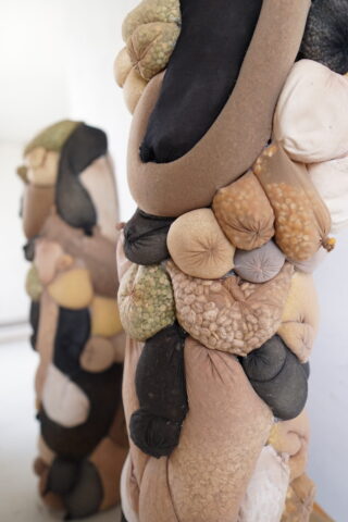 In einem weißen Raum sind zwei abstrakte Skulpturen aus Stoffbeuteln unterschiedlicher Form und Farbe ausgestellt, eine im Vordergrund, die andere unscharf im Hintergrund.