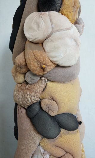 Eine Nahaufnahme verschiedener Stoffe und Textilien, die zu organischen, abgerundeten Formen in neutralen und gedämpften Tönen gestopft und genäht und vertikal vor einem schlichten Hintergrund angeordnet sind.