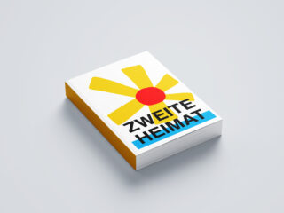 Auf einem hellgrauen Untergrund liegt ein Buch mit einer gelben Blume und roter Mitte auf dem Cover, auf dem in fetten schwarzen und blauen Lettern „ZWEITE HEIMAT“ steht.
