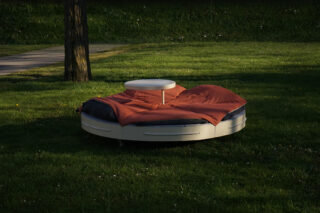 Ein rundes Bett mit roter Bettwäsche und einem runden Tisch in der Mitte steht im Freien auf einer Rasenfläche in der Nähe eines Baumes.