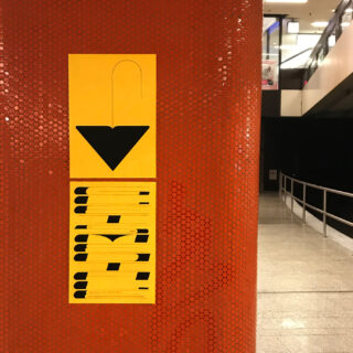 Ein gelbes Schild mit einem nach unten zeigenden schwarzen Pfeil ist an einer rot gefliesten Säule in einem öffentlichen Innenraum mit Geländer und Fliesenboden angebracht.